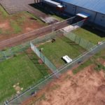 Aerial view of a fenced outdoor dog run with green grass, two dogs inside, and a white plastic play structure at a top-rated pet boarding service. The run is next to a large building with a blue roof and an adjacent parking lot. -Stay & Play Pet Resort